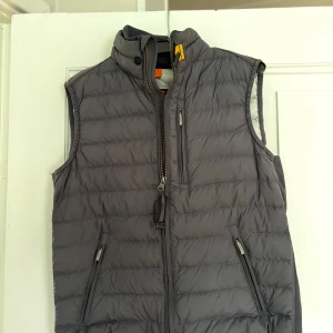 Grå dunväst från Parajumpers - Snygg grå dunväst från Parajumpers med quiltad design och hög krage. Västen har dragkedja framtill, tre fickor med dragkedja och en liten gul detalj vid halsen, Insidan är ljusgrå. Säljer den för den är för liten. Använd ett fåtal gånger så är som ny, hör av er vid frågor:)