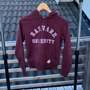 Vinröd Harvard hoodie från H&M XS - Snygg vinröd hoodie från H&M med Harvard University-tryck framtill. Tröjan har huva med snörning, magficka och långa ärmar. Perfekt för dig som gillar college-vibe och vill ha en bekväm och stilren look.