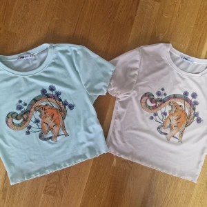 Pastellfärgad croppad t-shirt Fisher - Två croppade t-shirts från Fisher i pastellblått och ljusrosa med coolt tryck av tiger, orm och lila blommor. T-shirtsen har korta ärmar, rund hals och är gjorda i ett mjukt, stretchigt material som känns skönt mot huden.