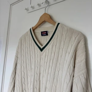 Kabelstickad v-ringad tröja Melbourne - Kabelstickad v-ringad tröja i beige från Melbourne Classix. Tröjan har gröna ränder vid halsringning, ärmslut och nederkant. Klassisk cricketstil med lång ärm och ribbade muddar. Perfekt för dig som gillar preppy vibes.