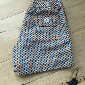 Mönstrade badshorts från Naive - Färgglada badshorts från Naive med blått, orange och vitt geometriskt mönster. Shortsen har elastisk midja med snörning och en bakficka med lock och logga. Perfekta för stranden eller poolen, riktigt sköna och lätta att bära.