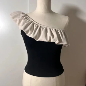 Svart offshoulder topp med volang - Snygg svart topp med offshoulder-design och stor beige volang som går diagonalt över bröstet. Toppen är tight och ärmlös, perfekt för dig som vill sticka ut med en cool och trendig look. Passar grymt till både jeans och kjol🩷🩷