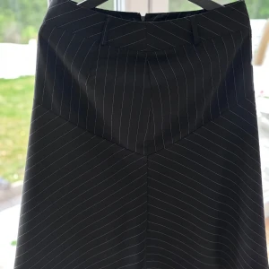 Svart randig långkjol med midjeband och silkesdetaljer - Fantastisk kvalité. Svart långkjol med tunna vita ränder och markerat midjeband. Kjolen har en lätt utsvängd form och bälteshällor i midjan. 