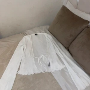 Vit omlottopp med knyt och volang - Superfin vit omlottopp med långa ärmar och knyt framtill. Tunt, lätt material med subtila ränder och volangkant nertill. Perfekt för lager-på-lager och ger en romantisk vibe till din outfit.