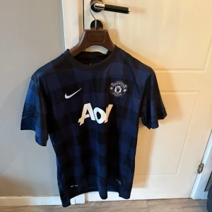 Manchester United rutiga bortatröja Nike - Snygg Manchester United bortatröja från Nike med rutigt mönster i mörkblått och svart. Tröjan har korta ärmar, rund hals och Aon-logga på bröstet. Tillverkad i lätt och ventilerande polyester, perfekt för match eller träning.