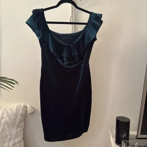 Mörkgrön sammetsklänning NLY One L - Supersnygg mörkgrön sammetsklänning från NLY One i storlek L. Upplever den dock som en M. Offshoulder-modell med volangdetaljer längs ringningen och figurnära passform. Perfekt för dig som vill sticka ut med en elegant look och mjukt material.