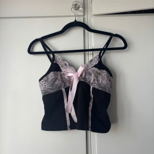 Svart linne med spets och rosett - Supersöt svart linne med tunna axelband, ljusrosa spetsdetaljer och en matchande rosett framtill, från SHEIN. Toppen har en croppad passform, perfekt för att lyfta vilken outfit som helst. Hör av dig om du vill ha fler bilder eller har några frågor! 