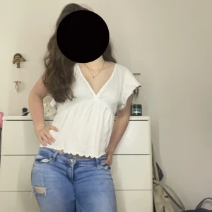 Vit blus med v-ringning och knyt - Säljer en luftig vit blus med v-ringning framtill och korta ärmar. Blusen har en söt knytning i ryggen och lätt volangkant nertill. Perfekt för varma dagar och enkel att styla med jeans eller kjol.