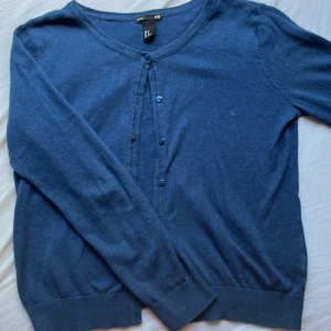 Blå kofta från H&M Basic, storlek M - En enkel och stilren blå kofta från H&M Basic i storlek M. Koftan har rund halsringning, långa ärmar och knäppning framtill med matchande blå knappar. Perfekt att slänga över en t-shirt eller linne för en chill look.
