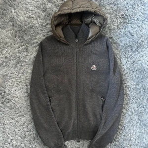 Grå Moncler pufferjacka XL / M - Sitter som M!!! Snygg grå pufferjacka från Moncler i storlek XL. Jackan har stickade ärmar och framsida i ull, medan rygg och huva är quiltade i glansig polyester. Dragkedja framtill och två fickor med dragkedja. Ikonisk Moncler-logga på bröstet.