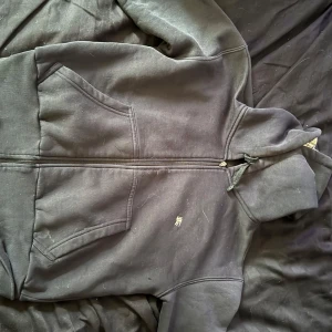 Svart Burberry hoodie med rutig huva - Svart hoodie från Burberry med dragkedja och klassisk rutig insida i huvan. Tröjan har två stora fickor framtill, snörning i huvan och en liten broderad logga på bröstet. Perfekt för dig som gillar stilrena och exklusiva streetwear-plagg.