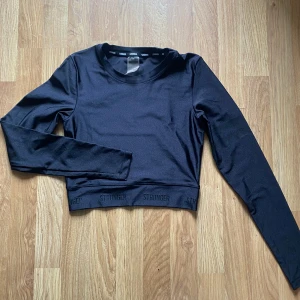 Svart croppad träningströja Stronger - Svart croppad långärmad träningströja från Stronger med diskreta ränder på ena ärmen och logga längs nederkanten. Tillverkad i stretchigt och glansigt material som sitter tight och är perfekt för gym eller yoga. Inga tydliga defekter. Hör av er vid frågor 💕