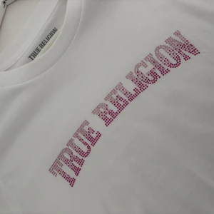 True Religion t-shirt - Vit t-shirt från True Religion med korta ärmar och rund hals. Framsidan har ett stort rosa paljettmönster med märkesloggan. Tyvärr har den aldrig kommit till användning. Köptes på RagLady i Helsingborg för 600kr. Den är i storlek L och sitter skönt och luftigt på mig som har Storlek M.