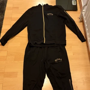 Svart tracksuit från Hugo Boss - Snygg svart tracksuit från Hugo Boss med gulddetaljer och logga på både jacka och byxa. Storlek xl passar L