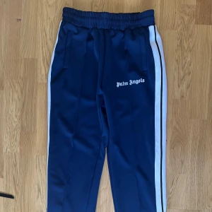 Blåa Palm Angels trackpants M - Säljer ett par blåa trackpants från Palm Angels med vita sidoränder och broderad logga på benet. Byxorna har elastisk midja och är gjorda i glansig polyester för en sportig vibe. Perfekta för dig som gillar streetwear och vill sticka ut.