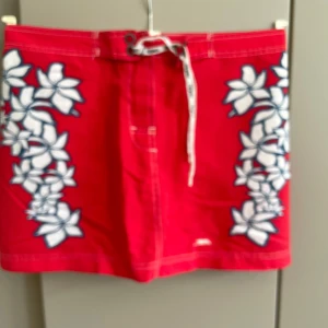 Röd  blommig kjol Trespass S - Kjol  från Trespass med vita blommor längs sidorna och snörning i midjan. Kjolen har två bakfickor, varav en med Trespass-logga. Perfekta för stranden eller poolen, tillverkade i snabbtorkande material. Midjemått 78/80 cm 