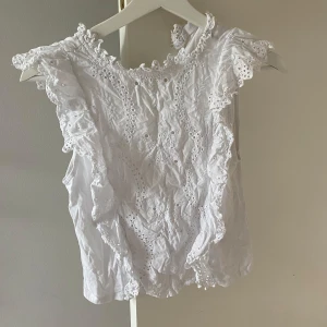 Vit blus med spets och volanger - Superfin vit blus i bomull med broderade spetsdetaljer och volanger längs axlarna. Ärmlös modell och rund halsringning. Perfekt för dig som gillar en söt och somrig stil!💗