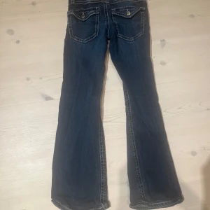 Mörkblå bootcut jeans med kontrastsömmar - Snygga mörkblå jeans med bootcut passform och markerade vita kontrastsömmar. Jeansen har klassiska fem fickor, varav bakfickorna har lock och knapp. Perfekta för dig som gillar en retroinspirerad look.