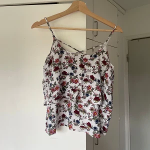 Blommig linnetopp med tunna axelband - Supersöt vit linnetopp med blommigt mönster i rött, rosa och blått. Toppen har tunna axelband och en luftig, flowy passform som är perfekt för varma dagar. Perfekt för dig som gillar romantiska vibes och somriga outfits.