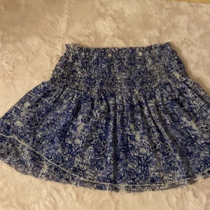 Blåvit mönstrad kjol Miss Milla - Supersöt kort kjol från Miss Milla i blått och vitt med ett unikt mönster. Kjolen har smockad midja och volangig nederdel som ger en luftig och skön känsla. Perfekt för dig som gillar en feminin och trendig stil.