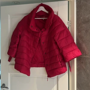 Röd pufferjacka med kort ärm - Snygg röd pufferjacka med korta ärmar och cool krage. Jackan har dragkedja och tryckknappar framtill samt banddetaljer vid ärmarna. Perfekt för dig som vill sticka ut med en färgstark look under kyliga dagar.