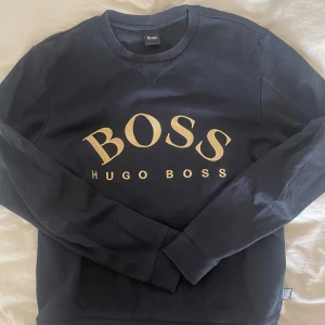 College-tröja Hugo Boss - College-tröja Hugo Boss svart med guldig text i storlek S. 