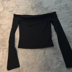 Svart offshoulder ribbad topp - Säljer en svart offshoulder topp med långa ärmar och ribbad struktur. Sitter tight och har en snygg croppad passform som framhäver axlarna. Perfekt för dig som gillar stilrena och trendiga plagg.