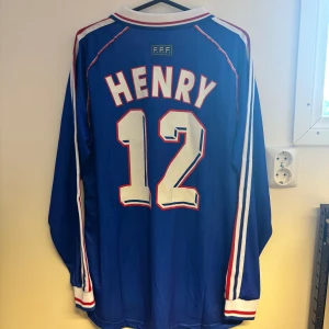 Frankrike VM 98 Henry #12 långärmad tröja - Legendarsik blå långärmad Frankrike-tröja från VM 1998 med Henry #12 på ryggen. Adidas-logga, broderad tupp och FIFA World Cup-detaljer. Röd och vit rand på krage och ärmslut, klassiska vita och röda ränder över bröstet. Tillverkad i ventilerande polyester.
