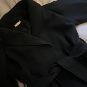 Svart kappa med bälte från Gina Tricot - Stilren svart kappa från Gina Tricot i storlek XS. Klassisk modell med bred krage och knytskärp i midjan för en snygg siluett. Perfekt för dig som gillar minimalistisk och tidlös. Perfekt nu till hösten, jag köpte den för 800kr och har använt den ungefär 3-4 gånger💞💞