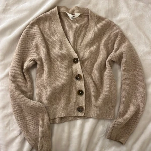 Beige stickad kofta i xxs - Säljer en beige stickad kofta från H&M med bruna knappar framtill. Koftan har lång ärm och en klassisk v-ringning. Perfekt att slänga över en t-shirt eller linne för en snygg och avslappnad look.