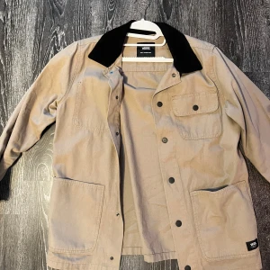 Beige overshirt från Vans med svart krage - Stilren beige overshirt från Vans med svart krage i kontrast, stora fickor framtill och knappar längs hela framsidan. Perfekt för lager-på-lager och chill streetstyle. Klassisk design med en bröstficka med lock och två stora sidofickor.