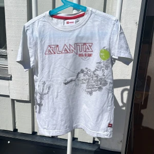 Vit LEGO Wear t-shirt Atlantis tryck - Vit t-shirt från LEGO Wear med coolt Atlantis-tryck i rött och grått framtill samt en grön detalj. Klassisk passform med rund hals och korta ärmar. Tillverkad i mjuk bomull, perfekt för dig som gillar äventyr och LEGO.