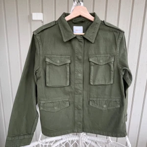 Olivgrön overshirt från Gina Tricot - Snygg olivgrön overshirt från Gina Tricot med fyra stora fickor framtill och axelklaffar. Jackan har klassisk krage, raka linjer och stängs med knappar. Perfekt lager-på-lager-plagg eller som jacka.