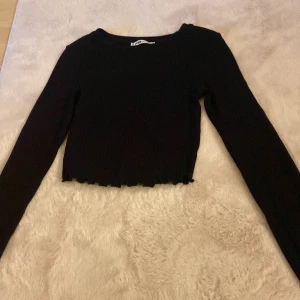 Svart croppad tröja från FB Sister - Svart ribbad långärmad tröja från FB Sister i croppad modell. Tröjan har en enkel rund halsringning och vågig kant nertill för en trendig look. Perfekt att matcha med höga jeans eller kjol.