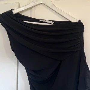 Svart oneshoulder topp från NA-KD - Elegant svart topp från NA-KD med en stilren oneshoulder-design. Klänningen har draperade detaljer som ger en sofistikerad look. Perfekt för en kväll ute eller en speciell tillställning. Endast använd 1 gång 