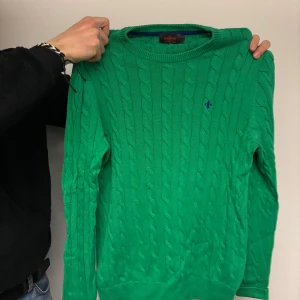 Grön kabelstickad tröja från Morris - Säljer en snygg grön kabelstickad tröja från Morris med deras ikoniska logga broderad på bröstet. Tröjan har långa ärmar och en rund halsringning. Perfekt för en stilren look!