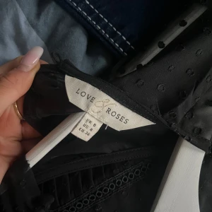 Svart blus från Love & Roses - Säljer en elegant svart blus från Love & Roses som är köpt i London. Blusen har korta puffärmar och fina spetsdetaljer på framsidan. Den är tillverkad i ett lätt och luftigt material med en diskret prickig struktur på ärmarna. 