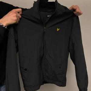 Svart vindjacka från Lyle & Scott - Säljer en stilren svart vindjacka från Lyle & Scott med dragkedja och broderad logga på bröstet. Jackan har långa ärmar och praktiska fickor framtill. Perfekt för blåsiga dagar!