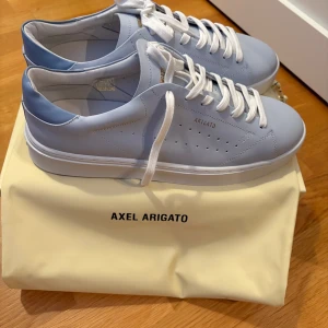 Vita sneakers från Axel Arigato - Snygga vita sneakers från Axel Arigato med stilren design och klassisk snörning. Skorna har en ljusblå detalj vid hälen och perforeringar på sidan för extra stil. Perfekta för en trendig och avslappnad look.