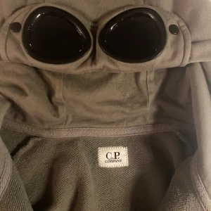 Grön jacka med glasögon från C.P. Company - Säljer en unik grön jacka från C.P. Company med inbyggda glasögon i huvan. Jackan har dragkedja framtill och är långärmad. Perfekt för dig som vill ha en stilren och funktionell look.