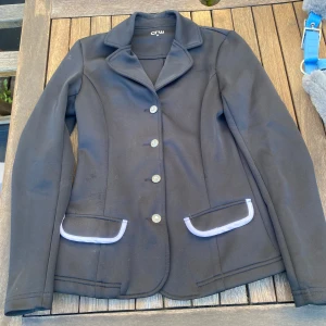 Svart kavaj från CRW - Svart ridkavaj med vita detaljer i storlek 34, något sliten men inte trasig.