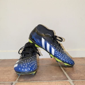 Blå och svarta fotbollsskor från Adidas(Predetor) - Snygga fotbollsskor i blått och svart med vita detaljer från Adidas. Skorna har en unik textur och snörning för optimal passform. Sulan är grön och designad för bra grepp på planen. Predator st 38 2/3 utan box och påse. Vi kan diskutera om pris!