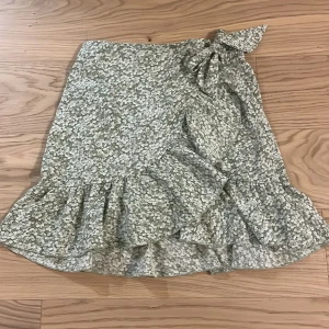 Blommig omlottkjol med volang - Söt kortkjol med blommigt mönster i grönt och vitt. Kjolen har en omlott-design med knytning i midjan och volangdetaljer längs nederkanten. Perfekt för en somrig look! Storlek: 152