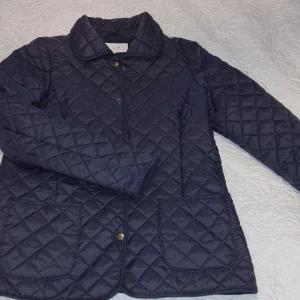 Mörkblå quiltad jacka  märke Vila,stM - Snygg mörkblå quiltad jacka med knappar framtill och två praktiska fickor. Jackan har en klassisk krage och långa ärmar, perfekt för kyligare dagar nu i övergångsperiod. En stilren design som passar till många tillfällen, mkt fint skick