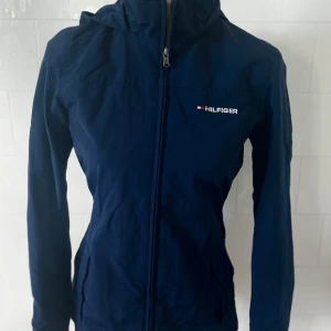 Mörkblå jacka från Hilfiger - Snygg mörkblå jacka från Hilfiger med dragkedja och huva. Huvan kan stoppas in i kragen. Jackan har långa ärmar och en bekväm passform. Tillverkad i polyester och nylon, perfekt för vår och höst. Storlek XXS. Använd fåtal gånger. I nyskick. 