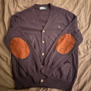 Blå kofta från Polo Ralph Lauren - Säljer en stilren Blå kofta från Polo Ralph Lauren med knappar och bruna mockadetaljer på armbågarna. Koftan har en v-ringad design och ribbade muddar vid ärmslut och nederkant. Perfekt för en klassisk look! Sitter mer som S. Har lagat några små hål