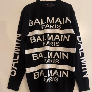 Svart och vit tröja från Balmain - Snygg svart tröja från Balmain med vit text 'BALMAIN PARIS' tryckt flera gånger över framsidan och ärmarna. Tröjan har en klassisk rund hals och långa ärmar. Perfekt för en stilren och modern look.