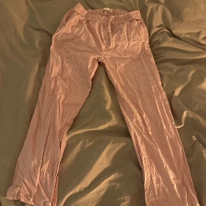 Randiga pyjamasbyxor - Säljer ett par bekväma randiga byxor i ljusrosa och vit. Byxorna har en elastisk midja med dragsko för justerbar passform. Perfekta för en avslappnad stil.