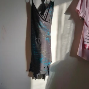 Svart glittrig klänning från Sisters Point - Snygg svart klänning från Sisters Point med glittriga detaljer och smala axelband. Perfekt för en kväll ute! Klänningen har en omlott-design och en kort längd som ger en trendig look.