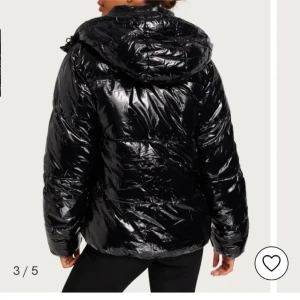 Svart glansig pufferjacka - Snygg svart pufferjacka med glansig finish och huva. Jackan har dragkedja framtill och långa ärmar. Perfekt för kyliga dagar när du vill hålla stilen.